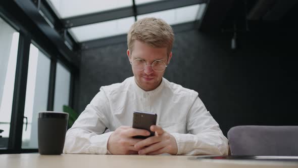 Handsome Man with Glasses is Using Smartphone in Cafe Reading News in Social Media Frontal Portrait