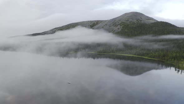 Aerial view of a low hanging clouds over a lake with small boat and mountain in the background.