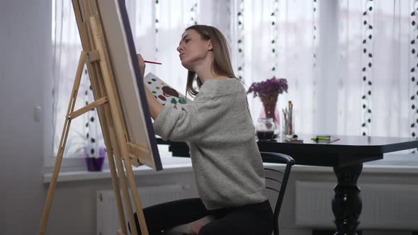 Side View Woman in Lotus Pose on Chair Taking Acrylic Paint with Brush Making Strokes on Canvas