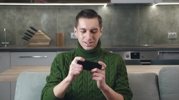 Young Man in Sweater Playing on His Smartphone Exciting Mobile Application Winning a Racing Game