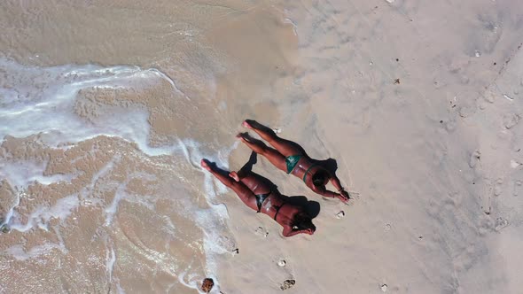 Two girls in bikini with tanned skin lying on white sand of exotic beach washed by white waves foami