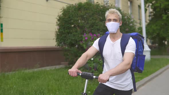 Young courier in medical mask delivering food with blue thermal backpack, riding a electric scooter