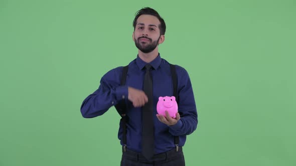 Happy Young Bearded Persian Businessman Holding Piggy Bank and Giving Thumbs Up