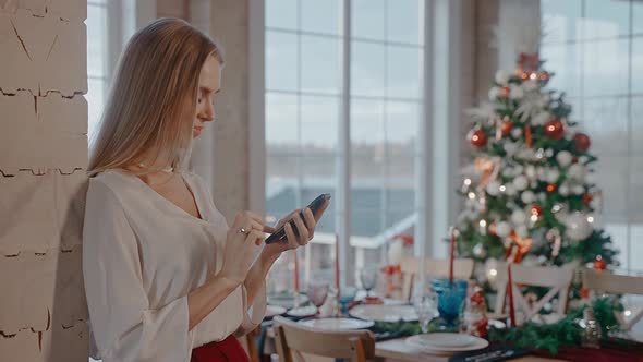 Blonde Woman is Texting on Phone in Front of Dining Table and Christmas Tree