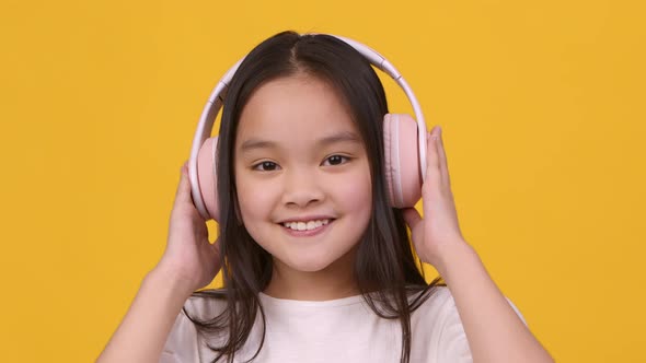 Portrait of Cute Little Asian Girl Wearing Big Wireless Headphones Smiling and Winking to Camera