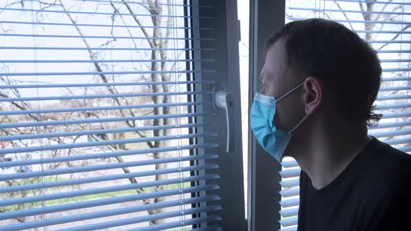 Young Man in Medical Mask Stands By the Window with Blinds and Sadly Looks Out Into the Street