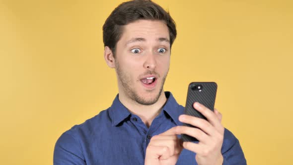 Casual Young Man Celebrating Success While Using Smartphone on Yellow Background