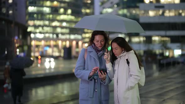 Two Women are Standing Under an Umbrella Looking at the Phone and Laughing Against the Background of