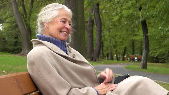 An Elderly Woman Sits on a Bench in a Park and Laughs