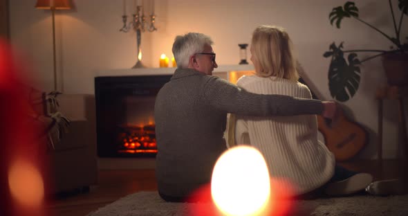 Back View of Aged Husband and Wife Hugging Relaxing on Carpet Near Fireplace