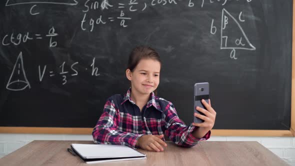 A Caucasian Schoolgirl Girl Sits at a Table Against the Background of a Chalk Board