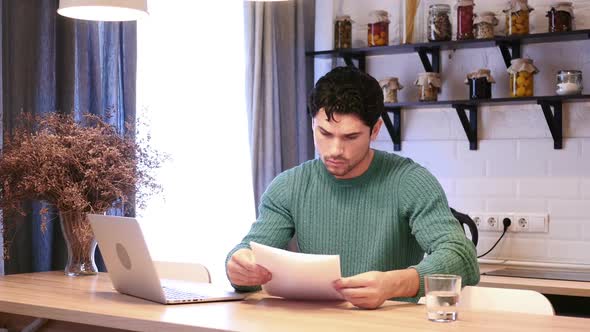 Man Working on Laptop and Reading Documents in Kitchen Paperwork