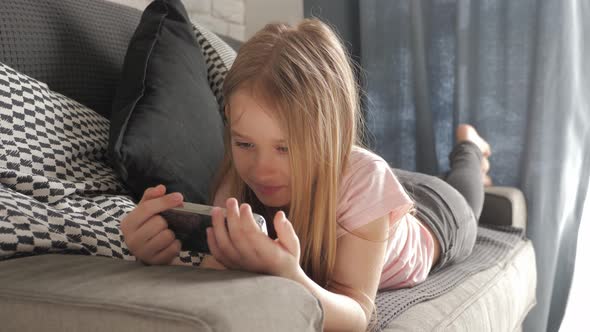 Little Girl Is Lying on the Bed and Looking Into Her Smartphone