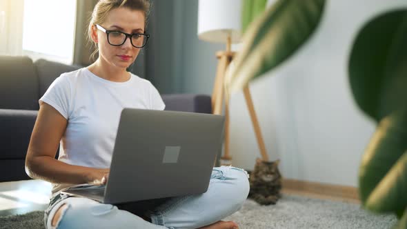 Woman with Glasses is Sitting on the Floor and Working on a Laptop