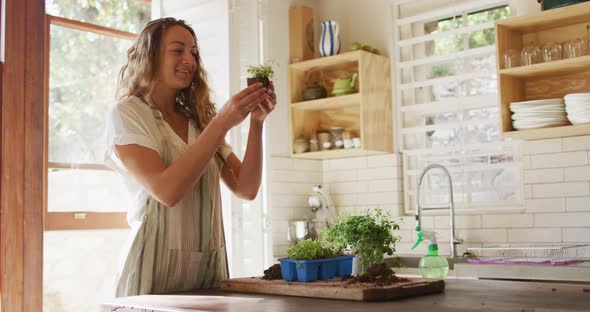 Smiling caucasian woman tending to potted plants standing in sunny cottage kitchen