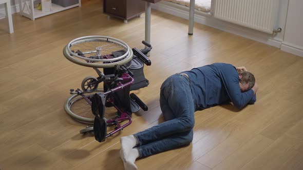 High Angle Wide Shot Desperate Crying Disabled Man Lying on Floor with Wheelchair Rolled Over