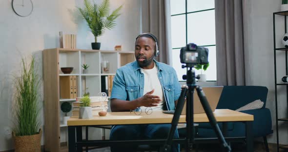 African American Blogger in Headset Streaming on Videocamera for Audience from Modern Apartment