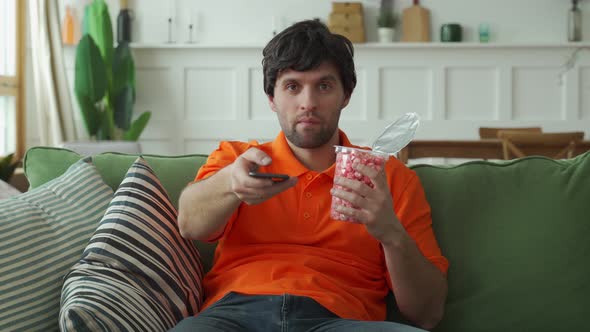 Man Sitting on Sofa Eating Popcorn Holding Remote Control and Switching Channels