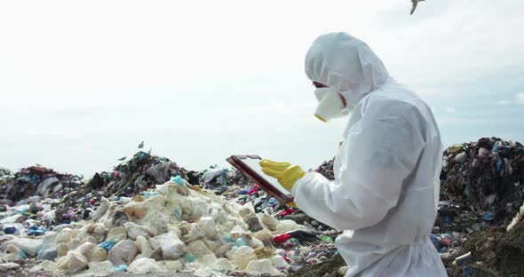 Man in Protective Suit and Gas Mask Noting in His Tablet at the Dump Landfill