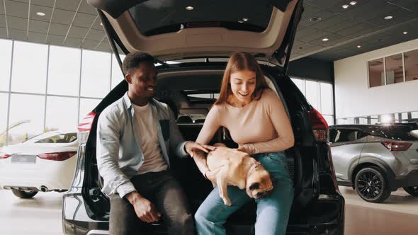 Smiling Multi Ethnic Couple Sitting on Open Trunk of Their New Luxury Car and