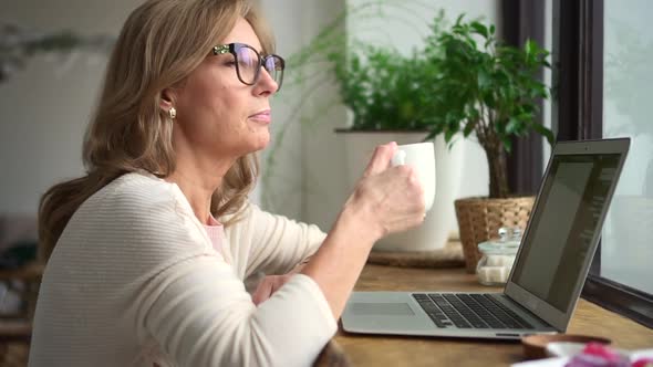 Woman Drinking Coffee and Working with Laptop at Table in Home Office