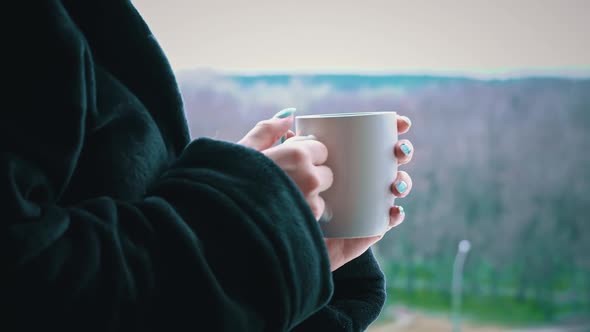 Woman Drinks Coffee From Cup in Black Bathrobe at an Open Window in the Morning