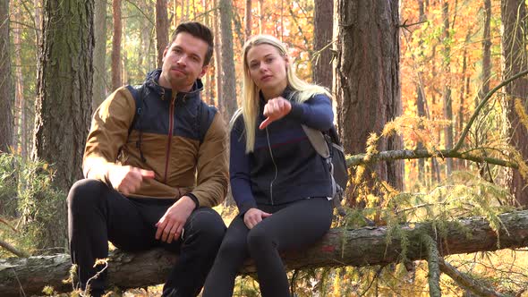 A Hiking Couple Sits on a Broken Tree in a Forest and Show a Thumb Down To the Camera