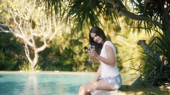 Woman Sits on Edge of Pool Splashes Water with Feet and Drinks Water From Glass