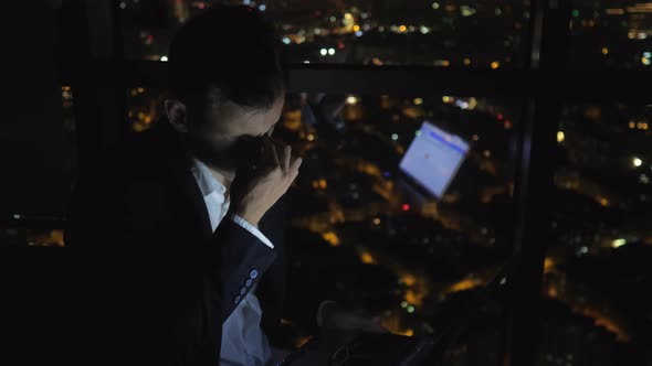 Tired Sleepy Man Is Working on His Computer at Night Near the Window with Cityscape.