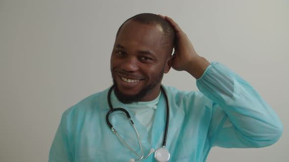 Portrait of Friendly Smiling Black Male Physician with Stethoscope Touching Head with Hand Indoors