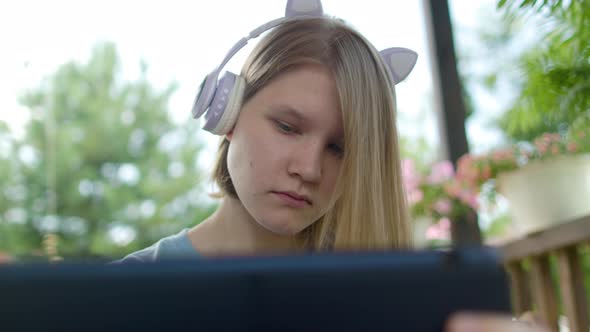 Teenage Girl Sits At Wooden Table In a Summer Cafe In Headphones With Phone