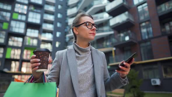 Woman Walking on a Business District Holds Takeaway Coffee Shopping Bags and Uses Smartphone