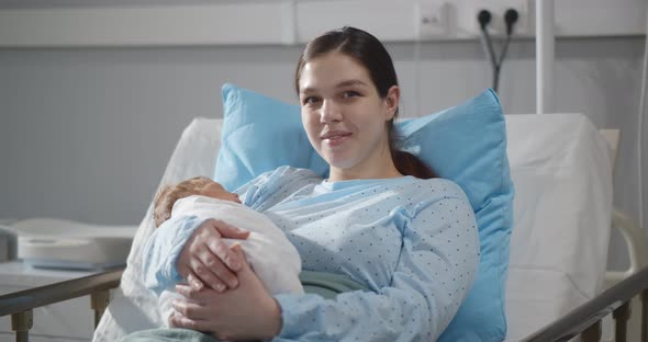 Happy Young Mother Sitting in Hospital Bed Holding Newborn Baby Swaddled in Blanket