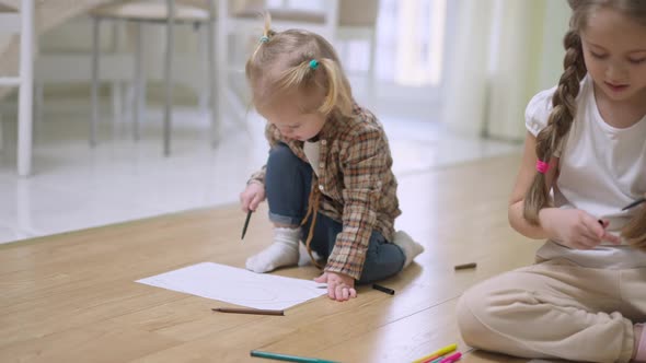 Cute Charming Little Blond Caucasian Girl Drawing Indoors with Blurred Brunette Sister