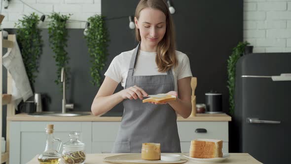A Woman Stands in an Apron in the Kitchen and Spreads Peanut Butter on Toast