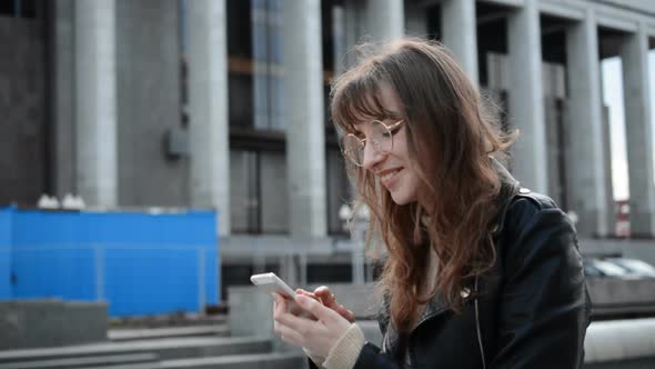 Portrait of Smiling Young Caucasian Woman Holding Smartphone and Reading Funny Message and Chatting