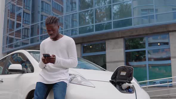 Stylish Young African American Man Standing Near an Electric Car Charging and Swiping in His