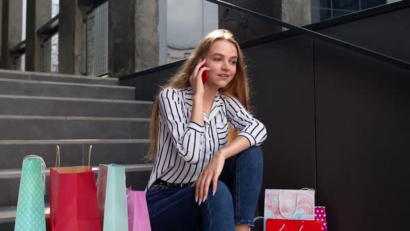 Girl Sitting on Stairs with Bags Talking on Mobile Phone About Sale in Shopping Mall in Black Friday