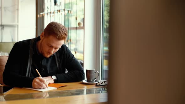 Portrait of Young Man Is Sitting in Cafe and Making Entries in His Notepad