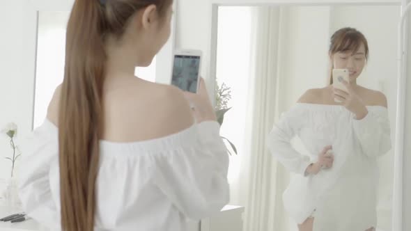 Beautiful young asian woman looking and selfie in the mirror with smartphone at living room.