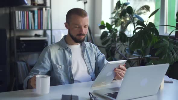 Young Man Using Tablet and Drinking Coffee While Sitting at Table in Office