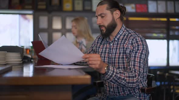 Side View Thoughtful Concentrated Man Analyzing Paperwork Sitting at Bar Counter with Blurred Woman