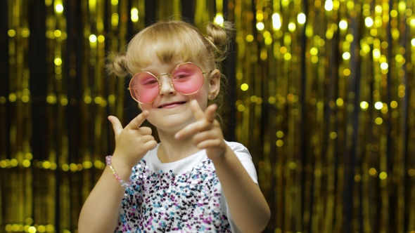 Child Smiling, Pointing Fingers at Camera. Girl Posing on Background with Foil Golden Curtain