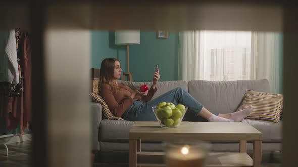 Beautiful Girl Eats a Red Apple Lying on Sofa and and Speaks Via Video Link