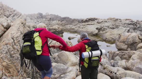 Senior hiker couple with backpacks and trekking poles holding each others hands while climbing rocks