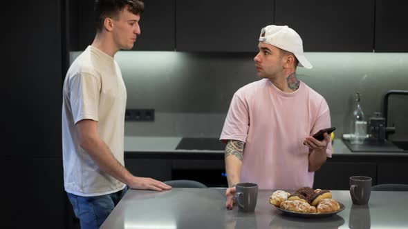 Young Gay Man Criticizing Tshirt of Partner Standing in Kitchen at Home