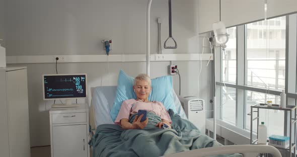 Smiling Senior Lady Having Video Call on Mobile Phone Lying in Hospital Bed