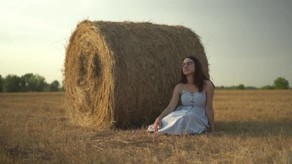 A Young Woman Sits Near a Haystack in a Field