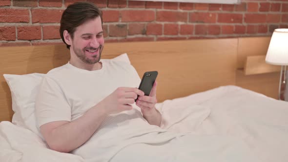 Casual Young Man Celebrating Success on Smartphone in Bed