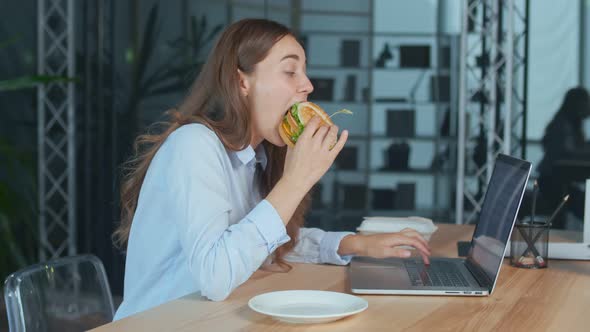 Young Businesswoman Eating Hamburger While Working on Laptop at the Office Table